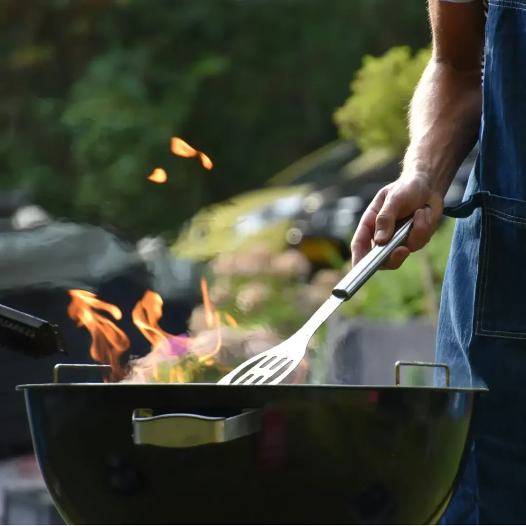 A person in a short-sleeve shirt and jeans is grilling outdoors, tending to a barbecue with a spatula over an open flame.
