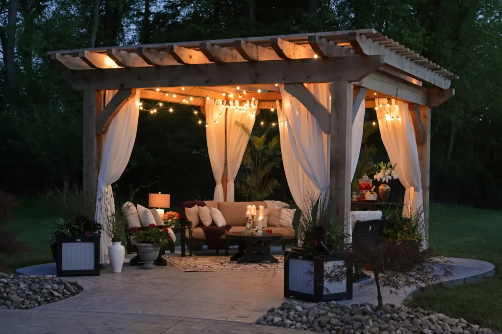 An elegantly lit wooden pergola adorned with sheer white curtains, string lights, and a cozy seating area with cushions and candles, set in a tranquil garden at dusk.