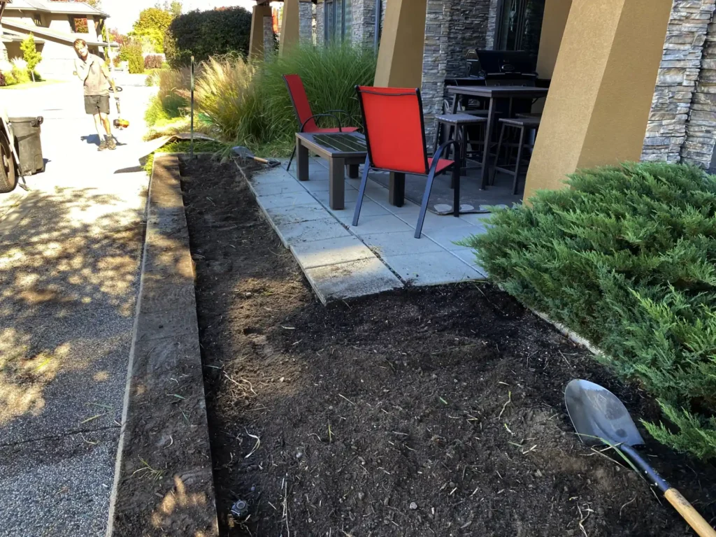 A freshly dug flower bed with a shovel lying on the soil in front of a patio area with two red chairs and a table. In the background, a person is walking on a sidewalk beside a lawn, carrying gardening tools.