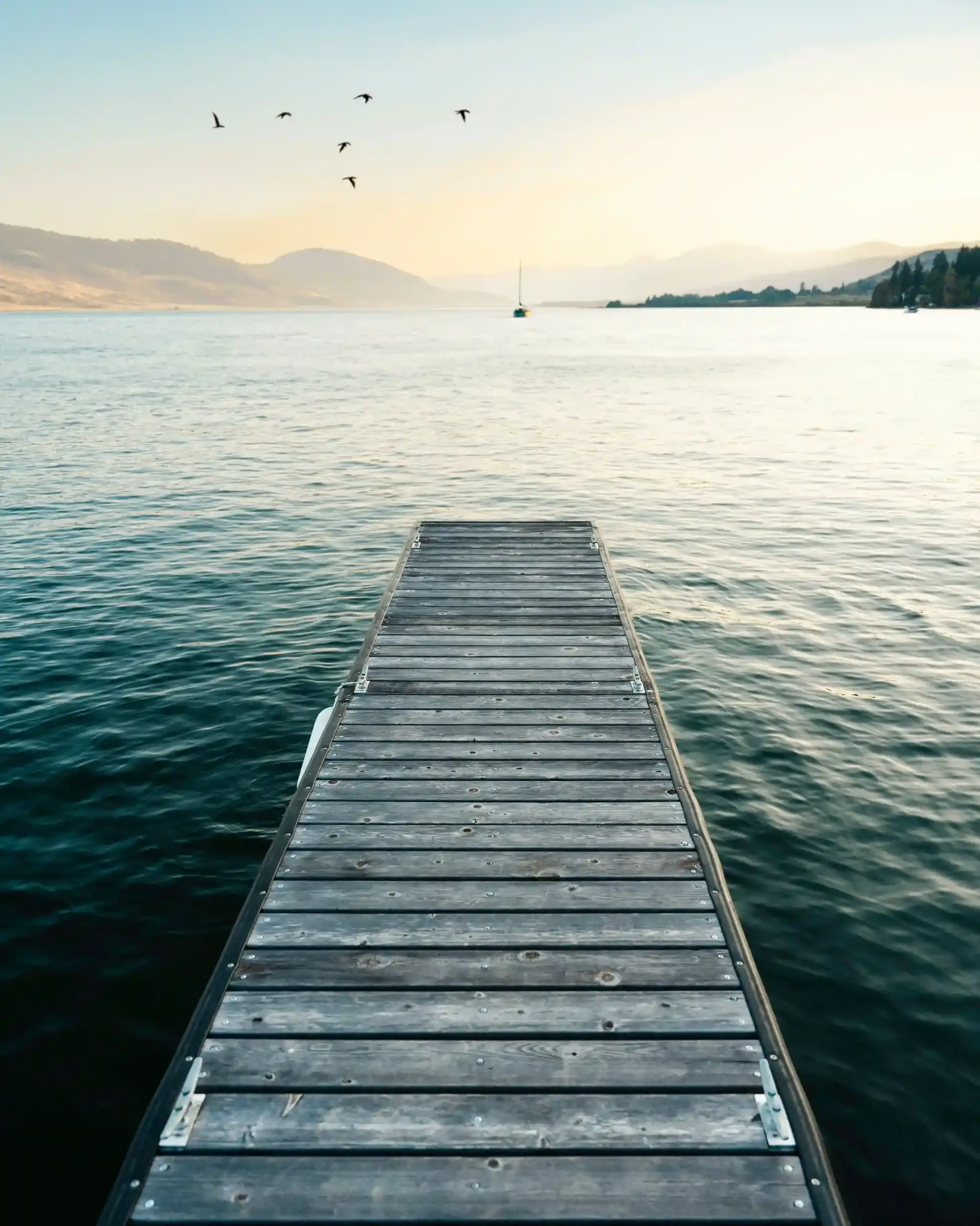 A wooden pier extending into calm waters, with hills in the distance, a sailboat to the right, and birds flying in the sky during early evening.