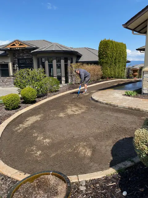 A person is raking soil in a curved garden bed in front of a residential house with a clear blue sky above.