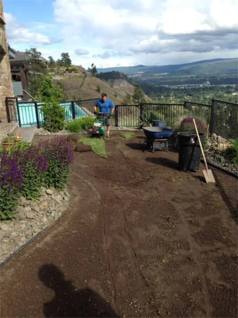 A person uses a rototiller on soil in a residential garden with a pool and house in the background, under a partly cloudy sky with a scenic view of distant hills.