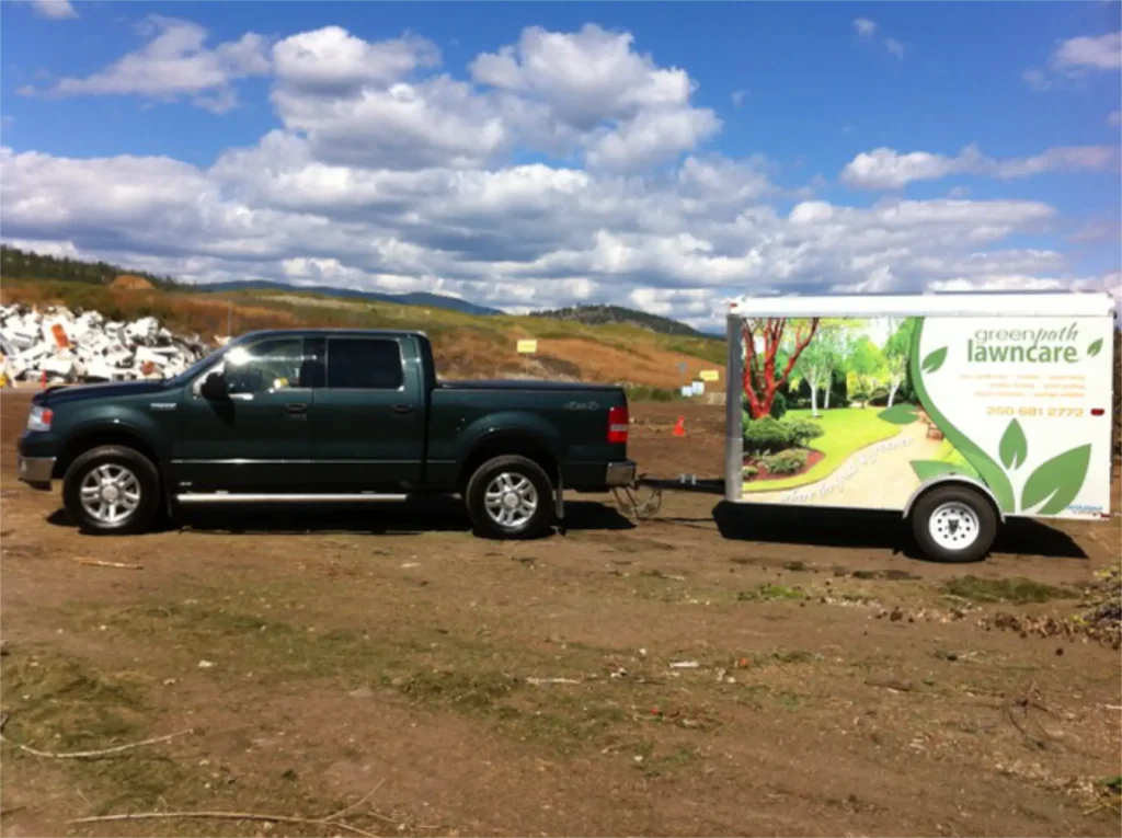 A green pickup truck is towing a white trailer adorned with a graphic advertising "Greenpath Lawncare," featuring a stylized image of a landscaped yard. The vehicle is parked on a dirt ground with a cluttered background of hills, clouds, and assorted debris.