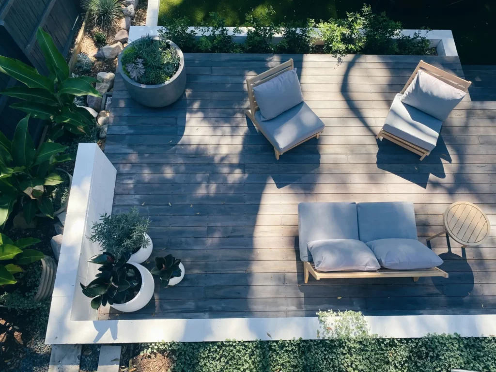 A modern outdoor deck with two single-seater wooden chairs with grey cushions, a double-seater sofa with similar cushions, a round wooden side table, and decorative potted plants. The deck is surrounded by a landscaped garden with a lush greenery border.