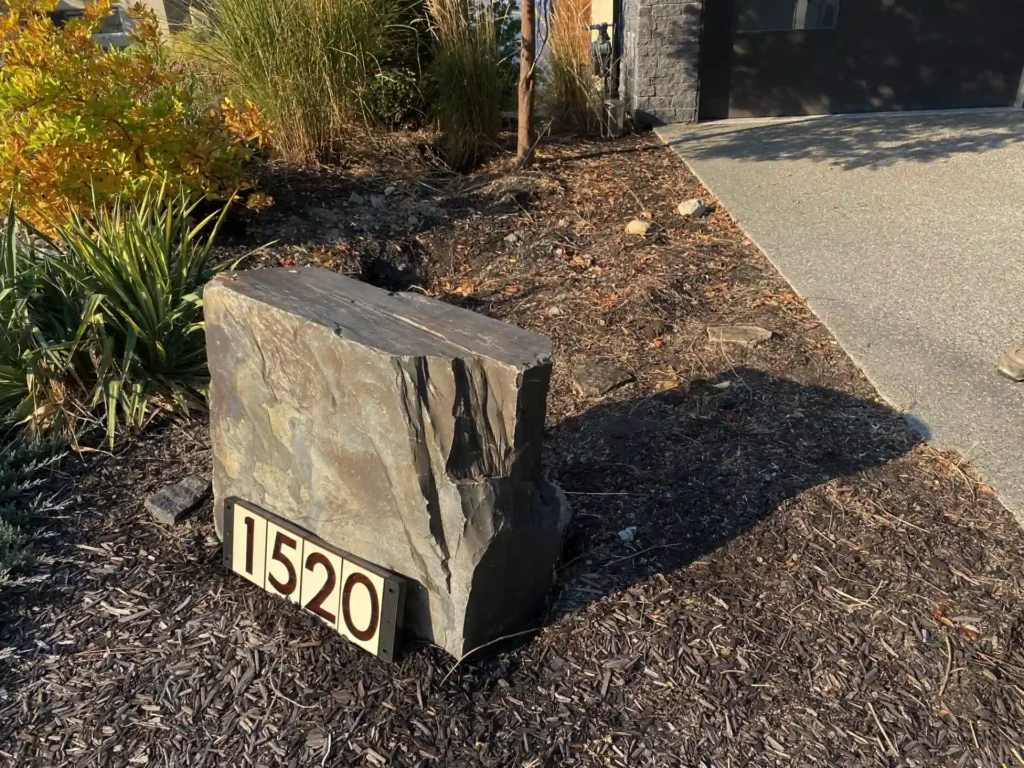 A large rectangular rock with the house number "1520" displayed on a plaque at its base, set in a landscaped area with mulch, shrubs, and a driveway to the right.