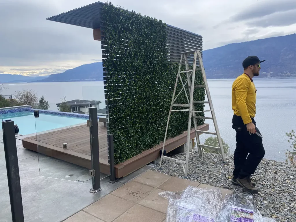 A person stands next to a vertical garden installation on a wooden structure with an adjacent infinity pool overlooking a calm body of water and mountains in the distance.