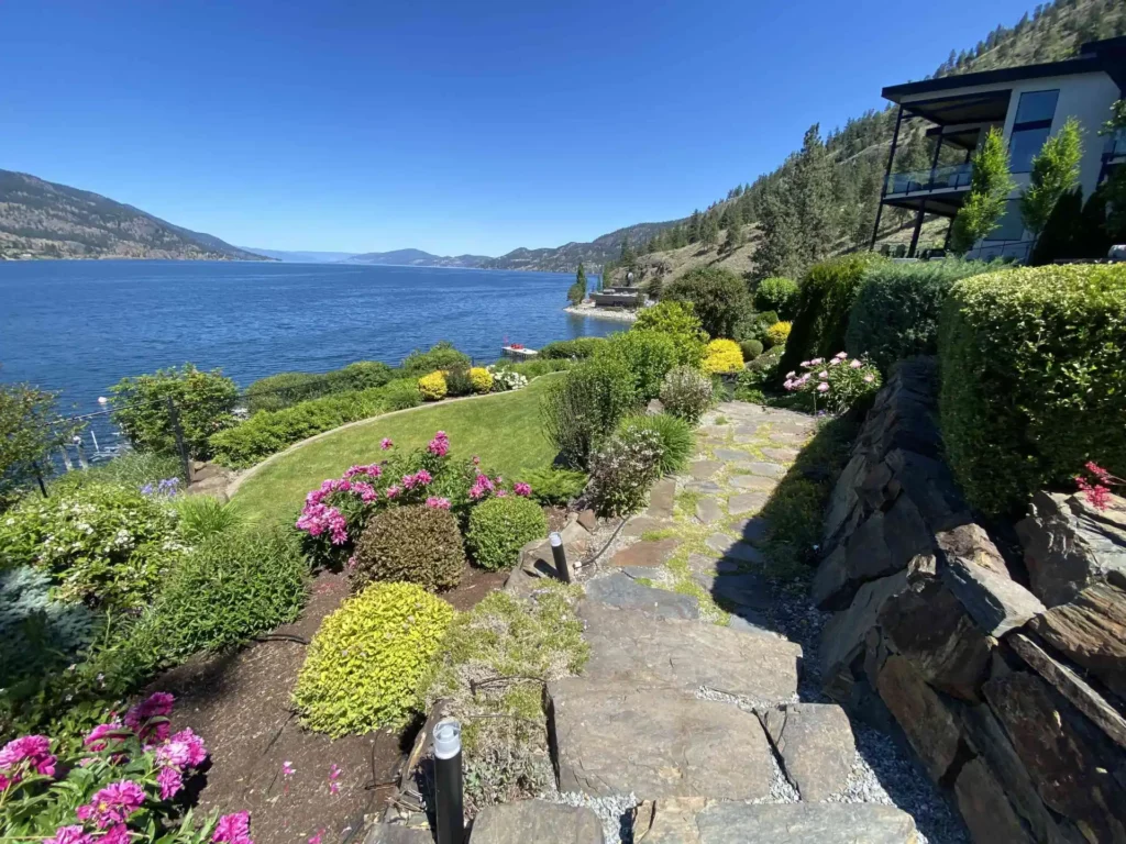 Stone pathway beside a lush garden leading towards a blue lake surrounded by hills with a clear sky above and part of a modern house visible on the right.