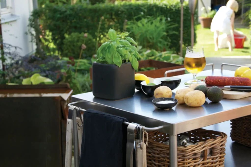 An outdoor cooking preparation area with fresh ingredients on a metal table: potatoes, avocados, a glass of beer, and a sausage. A potted basil plant is at the forefront, and in the blurred background, a person is tending to the garden.