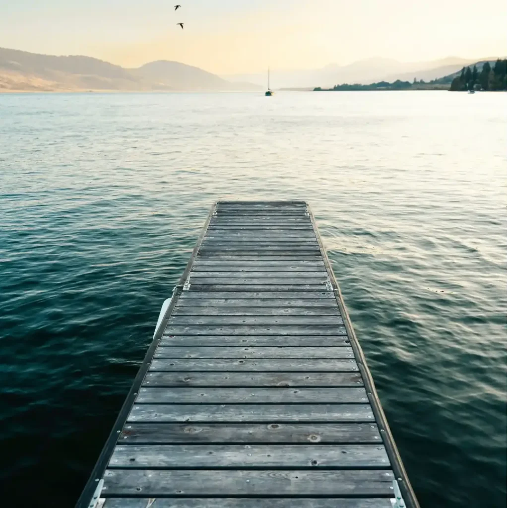 A wooden pier extends into a calm lake with mountains in the background and a few birds flying in the sky. A single sailboat is visible in the distance on the right. The lighting suggests either dawn or dusk.