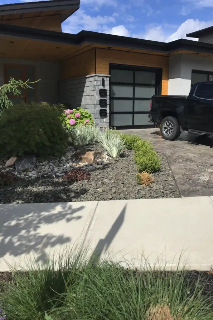 A modern house with a large black garage door, wooden siding, and a landscaped front yard with a variety of plants and rocks, partially obscured by a black pickup truck parked in the driveway.