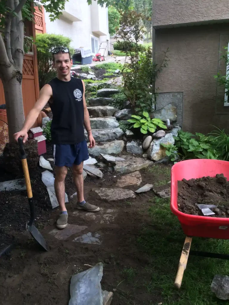 A man stands smiling by a stone pathway holding a shovel, with a wheelbarrow filled with dirt adjacent to him, suggesting garden work or landscaping.