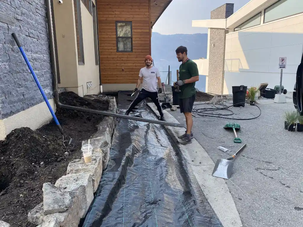 Two men working on landscaping a garden bed next to a building with soil and landscaping fabric, with some gardening tools and plants visible.