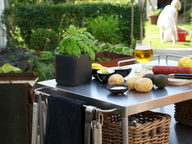 An outdoor cooking preparation area with a basil plant, freshly washed potatoes, half an avocado, a glass of beer, and other ingredients, with a child playing in the sunny garden in the background.