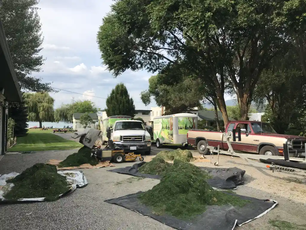 A residential driveway with landscaping work in progress, featuring trucks, a trailer, piles of debris, equipment, and tarps scattered around, with a lake and trees in the background.