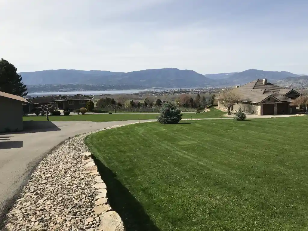 A scenic view of a suburban area with well-manicured lawns, single-story homes, and a backdrop of mountains and a lake under a clear sky.