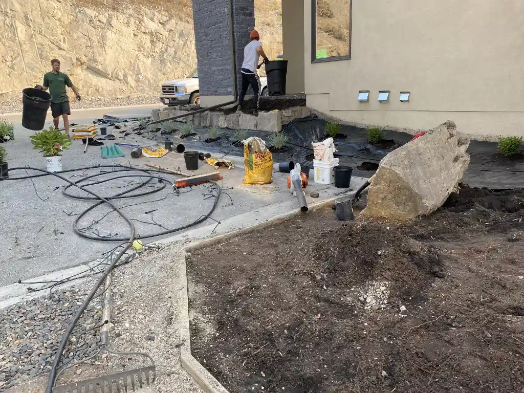 A landscaping project in progress outside a building, with garden tools, plants, soil, a large rock, and cables scattered around; two people are working in the area.