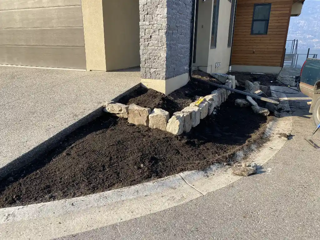 A garden bed in the process of being built or renovated in front of a house, featuring a pile of soil and a partial stone retaining wall on a sunny day.