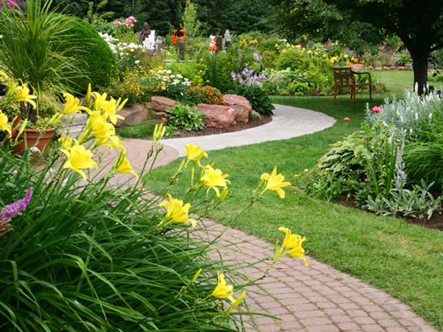 An image of a well-maintained garden with a winding path, featuring yellow lilies in the foreground and a variety of colorful flowers and plants throughout, with a wooden bench in the background.