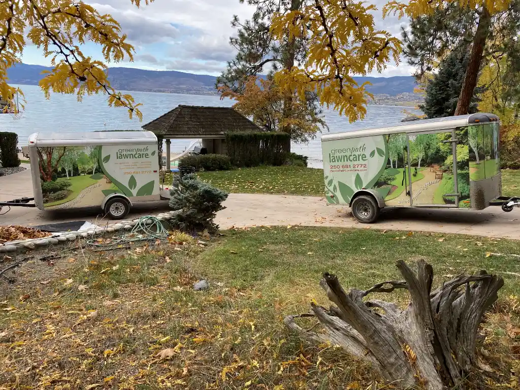 Two lawn care trailers with "greenpath lawncare" graphics parked on a driveway with a view of a lake and mountains in the background, framed by autumn leaves.