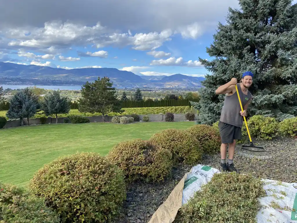 A smiling man holding a leaf rake stands on a tarp in a landscaped garden with trimmed hedges in the foreground and a scenic view of hills and a lake in the background under a partly cloudy sky.