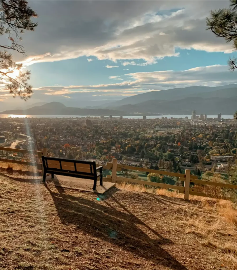 A solitary bench overlooking a panoramic view of a city with lush trees and a large body of water, backlit by a warm sunset with clouds scattered across the sky.