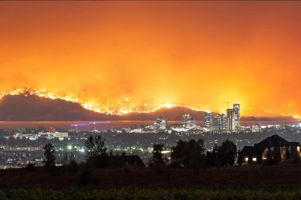 A night-time view of a city skyline with a large wildfire visible on a hillside in the background, illuminating the sky in a dramatic orange glow.